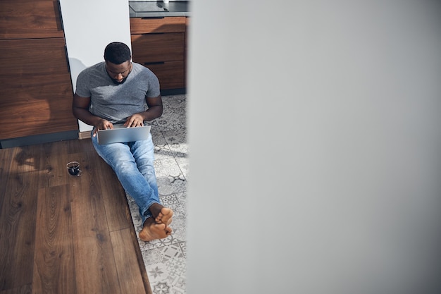 Concentrated international man sitting on the floor and looking at screen of his gadget while chatting online