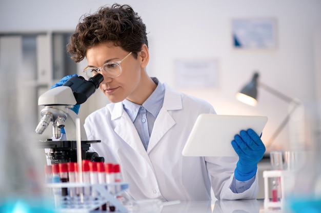 Concentrated laboratory technician using data on tablet while analyzing sample via microscope