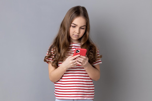 Concentrated little girl wearing striped Tshirt using smart phone looking at device screen