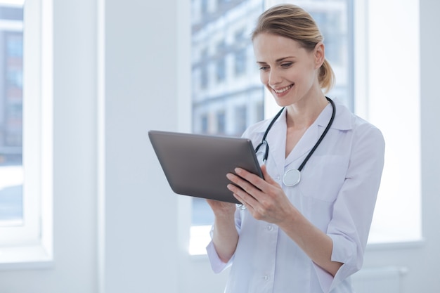 Concentrated lively professional practitioner enjoying working hours in the hospital while expressing interest and using tablet