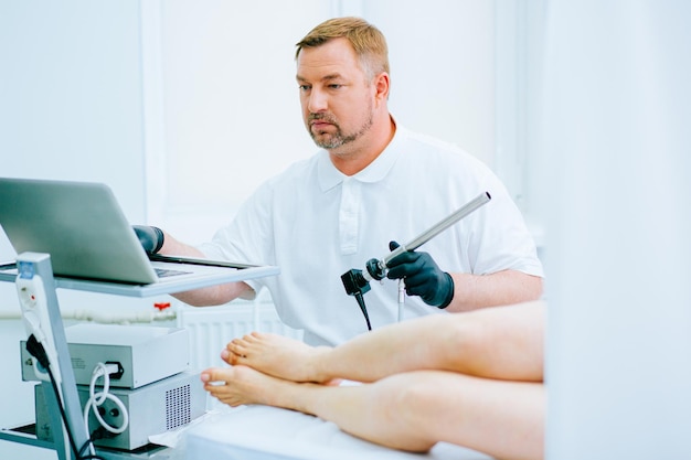 Concentrated male doctor looking at the screen while holding a video proctoscope. Legs of the patient near