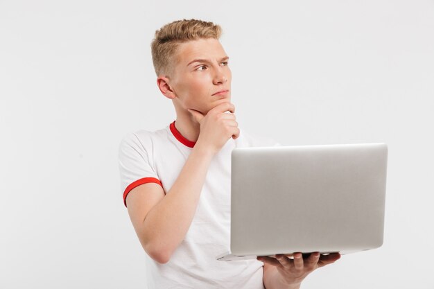 concentrated man 16-18 years old in t-shirt touching his chin and thinking while holding open laptop on hand isolated on white