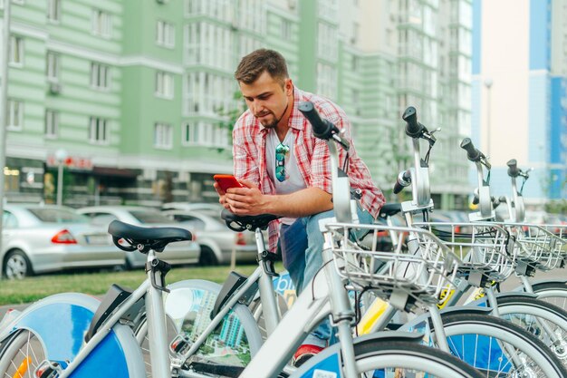 カジュアルな服を着た集中男性が自転車の列の近くに立ってスマートフォンで番組を共有しているひげを生やした男がレンタル自転車のそばに立ってスマートフォンを使用している