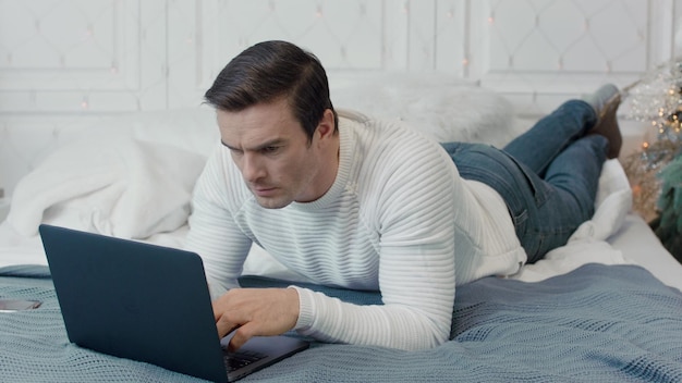 Concentrated man laying with computer on bed in luxury house