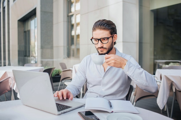 Concentrated man sits at table and works on laptop. He hold cup of tea on hand. There are opened notebook and pen at table. Businessman works.