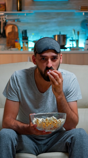 Uomo concentrato con la maschera per gli occhi del sonno seduto davanti alla televisione