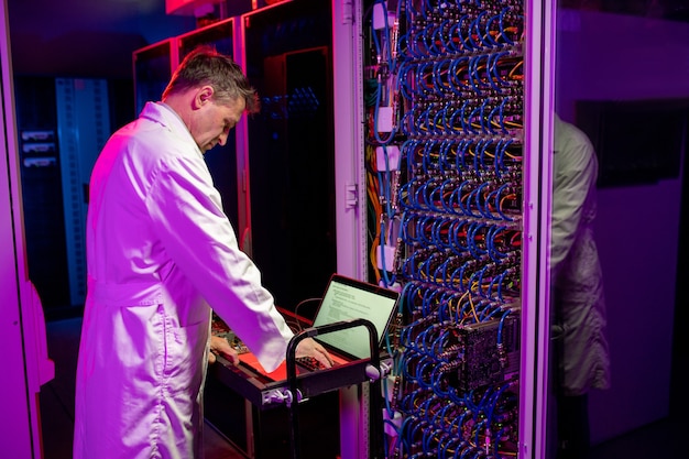 Concentrated mature network engineer in white coat standing at cart with laptop and configurating blade server in neon illuminated room