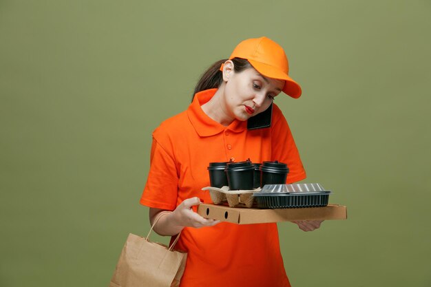 Concentrated middleaged delivery woman wearing uniform and cap holding pizza package food container egg tray plastic coffee cups and paper bag looking down talking on phone