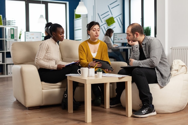 Concentrated multiethnic businesspeople reading annual financial report from tablet sitting on couch in modern startup business office