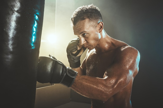 Concentrated muscular man is training actively and hard, hitting a punching bag.