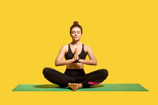 Сконцентрированная мирная женщина с пучком волос в обтягивающей спортивной одежде сидит на коврике, практикует йогу, держится за руки в жесте намасте и медитирует, расслабляется. студийный снимок на желтом фоне