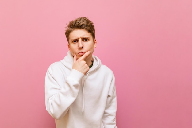 Concentrated pensive guy in white casual clothes stands on a pink background looking in camera