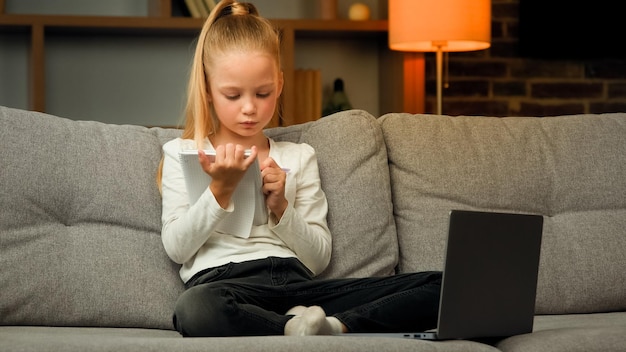 Concentrated pupil girl making homework at copybook on cosy sofa writing math tasks of elementary