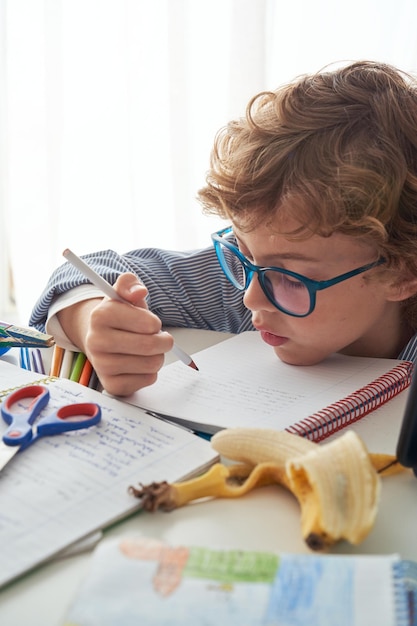 Concentrated schoolboy in eyeglasses with curly hair sitting at desk and doing assignment while studying at home