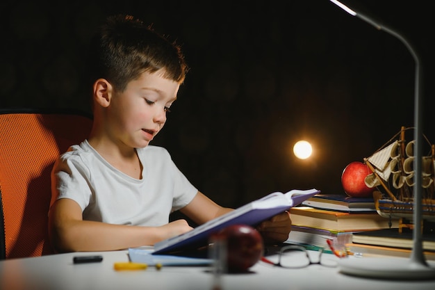 Concentrated schoolboy reading book at table with books, plant, lamp, colour pencils, apple, and textbook