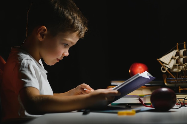 Сосредоточенный школьник читает книгу за столом с книгами, растением, лампой, цветными карандашами, яблоком и учебником