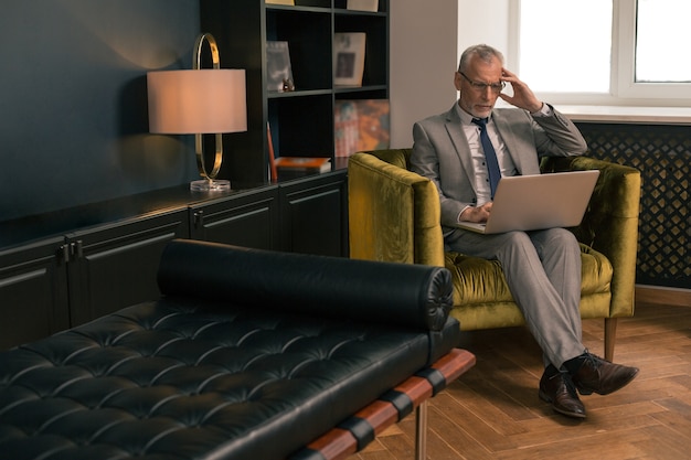 Concentrated serious gray-haired man sitting in an armchair indoors while working on his laptop