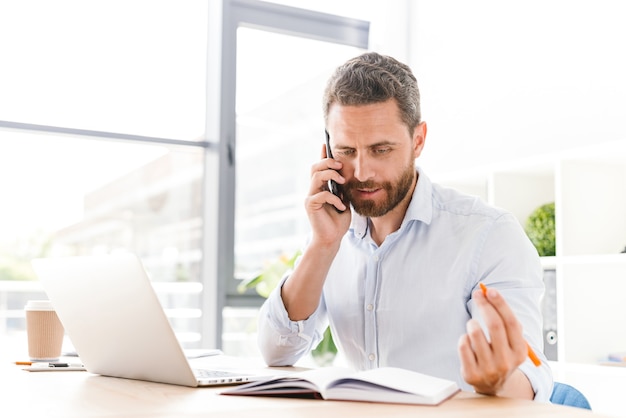 Foto uomo bello serio concentrato che parla dal telefono cellulare mentre si utilizza il computer portatile.