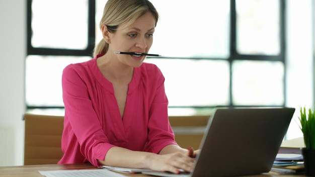 Concentrated serious woman looks at computer screens and holds pencil in mouth