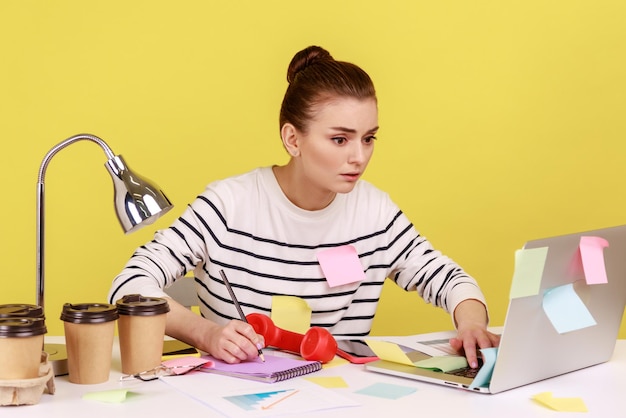 Concentrated serious woman office employee working on laptop and writing down information in notepad