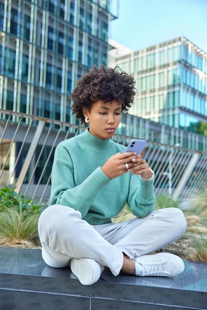 Concentrated serious young woman sits in lotus pose sends text messages and reads information on mobile phone outdoors