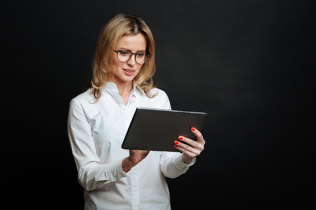 Сосредоточенная умная привлекательная женщина выражает восторг и использует планшет, стоя изолированно в черной стене