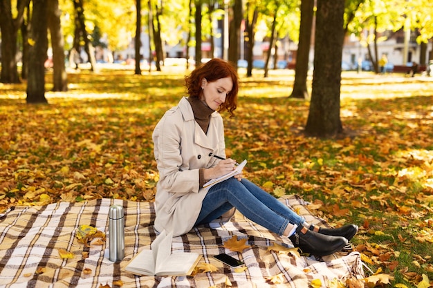 Concentrated smart young european redhaired lady student in\
raincoat sit with phone makes notes in diary