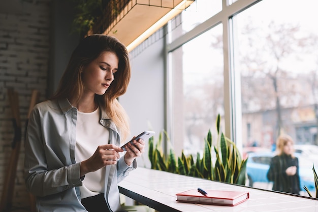 Concentrated teenage hipster girl sitting indoors and using mobile phone for blogging