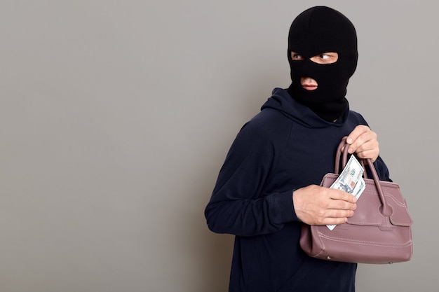 Concentrated thief dresses in balaclava and black turtleneck looks back and holds money and stolen woman's bag in his hands