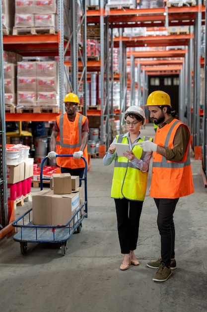 Concentrated warehouse coworkers in hardhats searching for items via online catalog on tablet while mover pushing storage cart