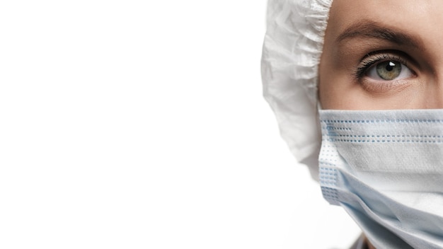 Concentrated woman doctor in surgical mask and cap is looking at camera, white background. Operation, medical practitioner, surgery, transplant, healthcare, medicine, COVID-19 concept. Close-up view