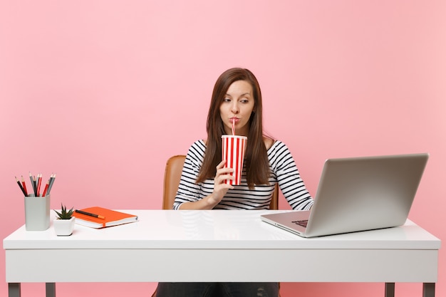 Donna concentrata che beve dalla tazza plactic con cola soda che lavora al progetto in ufficio alla scrivania bianca con laptop pc contemporaneo