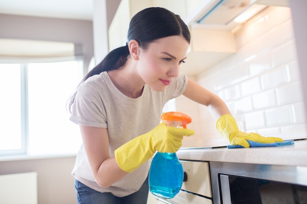 Concentrated woman stands in front of stove and look at it. She is cleaning the surface. Girl is doing that careful. She wears yellow gloves.