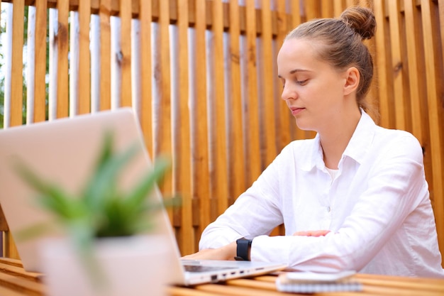 Concentrated woman wearing white shirt sitting in outdoor cafe working on laptop watching webinar online learning freelance job networking browsing
