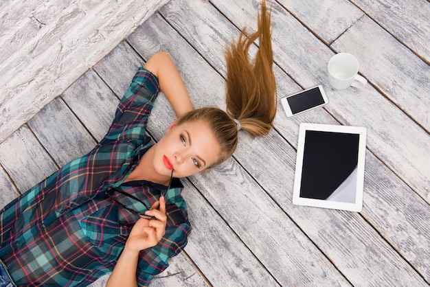 Concentrated woman with tablet, spectacles, phone and cup lying on the floar