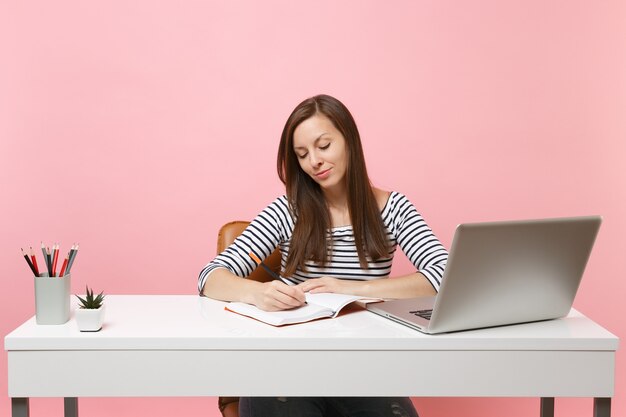 Donna concentrata che scrive note sul taccuino si siede, lavora alla scrivania bianca con un computer portatile contemporaneo