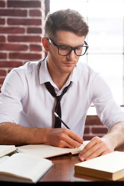 Concentrated on work. Confident young man in shirt and tie writing something in note pad while sitting at his working place