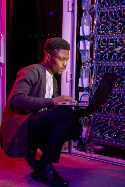 Сосредоточенный молодой афро-американский ИТ-инженер, присевший у серверного шкафа и анализирующий работу сервера с помощью портативного компьютера