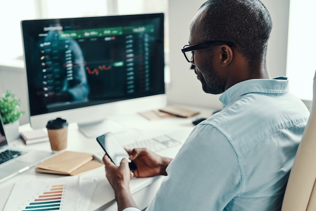 Concentrated young African man in shirt using smart phone while working in the office