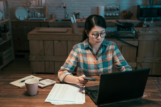 concentrated young asian chinese wife paying taxes through network on laptop computer. beautiful lady sitting at desk in home kitchen and using notebook pc at night with credit card in hand