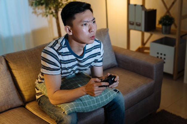 Concentrated young asian korean guy enjoying video game in dark\
house living room at late night. man playing with joystick sitting\
on couch. frowning face of male gamer relax in evening having\
fun.