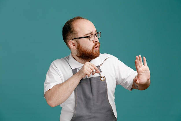 Concentrated young barber wearing uniform and glasses holding scissors keeping hand in air looking at his hand isolated on blue background