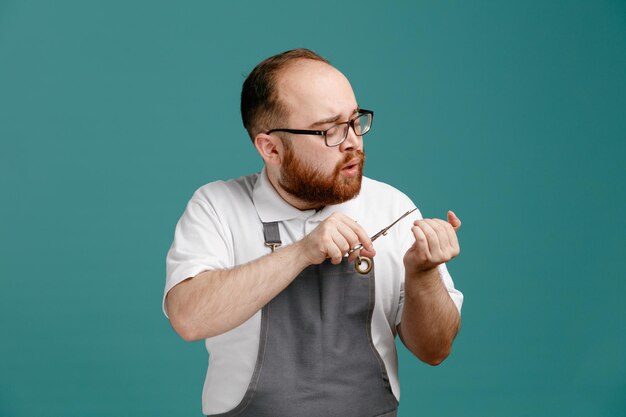 Concentrated young barber wearing uniform and glasses looking at his nails cutting nails with scissors isolated on blue background