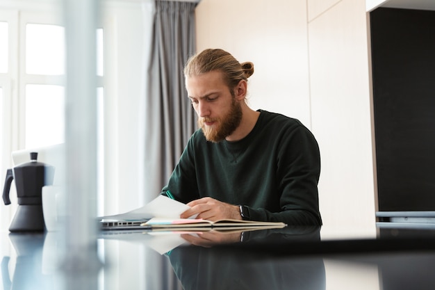 Photo concentrated young bearded man sitting in home using laptop computer work with documents.