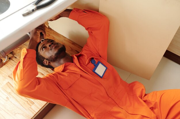 Concentrated young Black bearded plumber with badge on chest lying under sink and fixing clogged drains