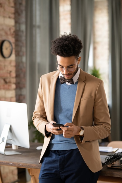 Concentrated young Black man in bow tie standing at office table and checking phone messages