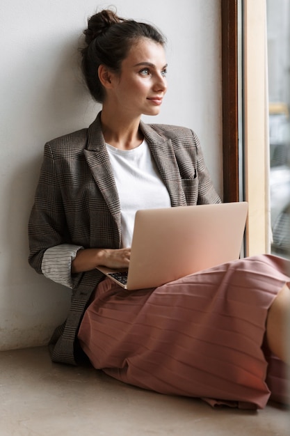 концентрированная молодая деловая женщина в помещении дома работает с портативным компьютером возле окна.