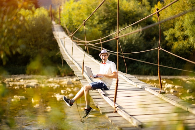 Concentrated young businessman in eyewear sit on bridge across river with laptop