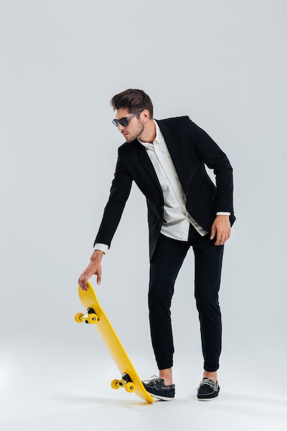 Concentrated young businessman in sunglasses and black suit going to ride a skateboard over gray wall