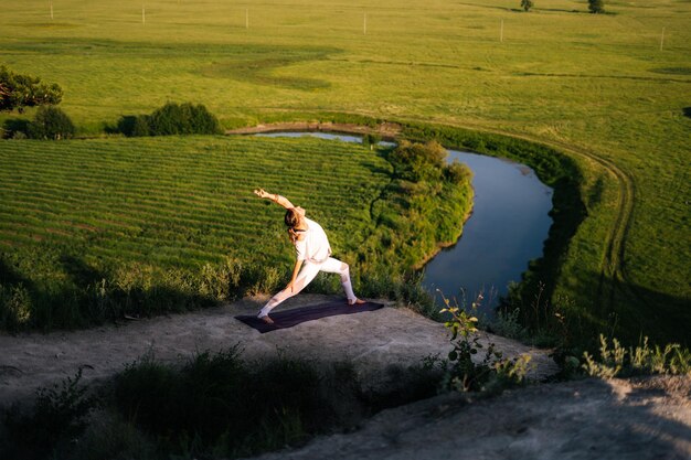 Concentrated young Caucasian woman is performing warrior yoga asana pose on top of rock background of flowing river and green meadow on summer sunny day Girl practicing Virabhadrasana